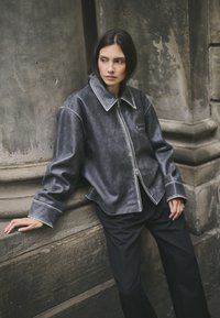 Grijze leren jas met een brede kraag, ritssluiting en witte stikselaccenten, gecombineerd met zwarte broek tegen een stenen achtergrond.