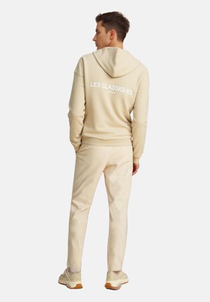 Beige hooded sweatshirt with white text on the back, paired with light beige trousers and sneakers, viewed from the back.