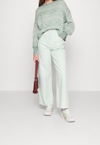 Hellgrüner gestrickter Pullover mit einem gemusterten Design, kombiniert mit mintgrünen weit geschnittenen Hosen. Das Model hält eine burgunderfarbene Tasche und trägt weiße Sneaker.