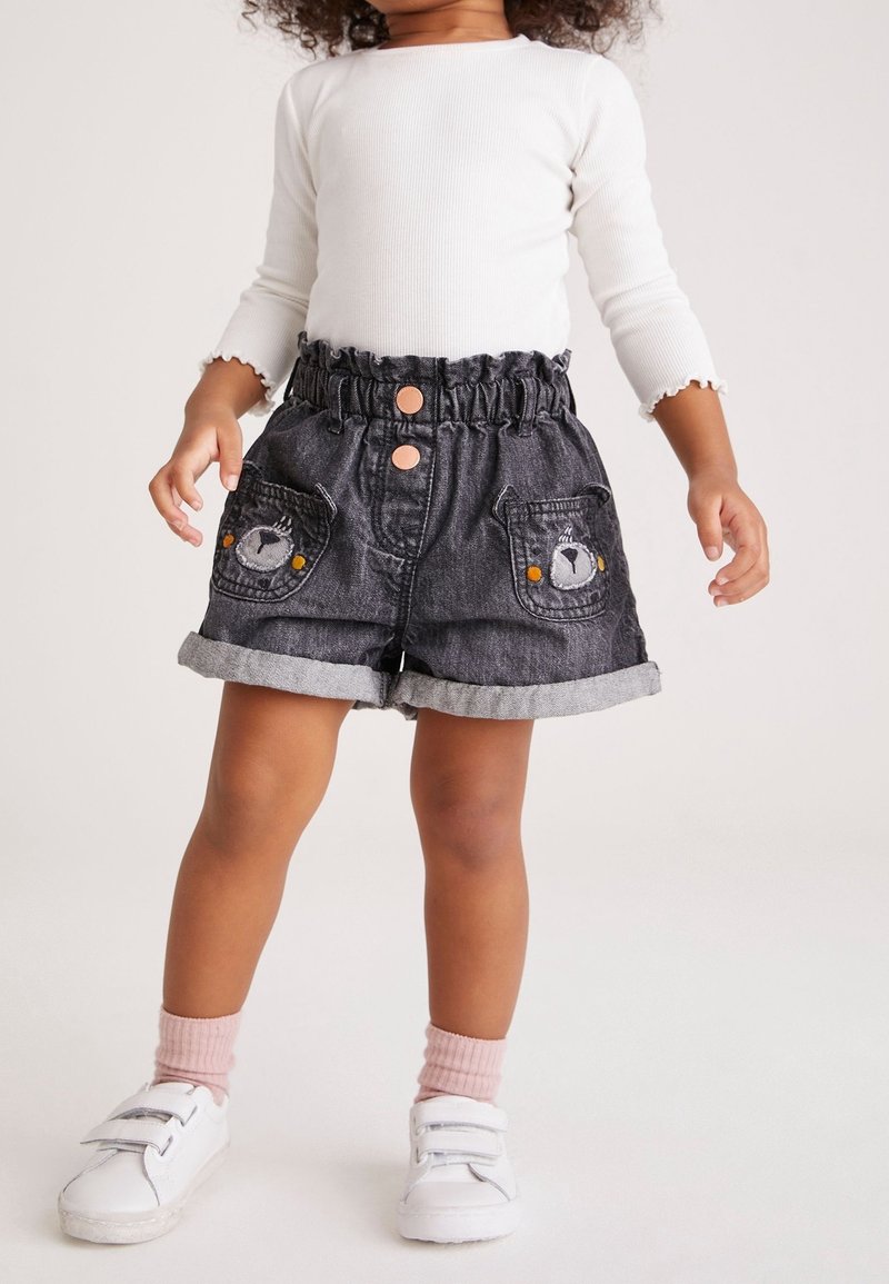 Tout-petit portant un haut blanc à manches longues, un short noir à ourlet roulé avec des écussons d’ours sur les poches, des chaussettes roses et des baskets blanches.