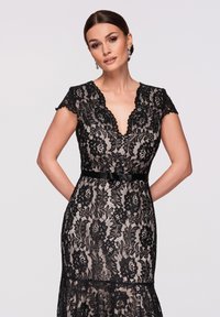 Femme portant une robe en dentelle noire à fleurs avec des manches courtes et un décolleté en V, debout les mains derrière le dos devant un fond uni.