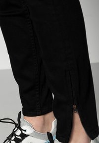 Pantalones de mezclilla negros con detalle de cremallera lateral, combinados con zapatillas blancas y grises que tienen cordones negros y acentos en azul claro.