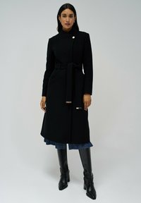Zwarte wollenjas met een ceintuur in de taille, hoge kraag en zijzakken. Bevat een opvallende zilveren knop en een soepele textuur.