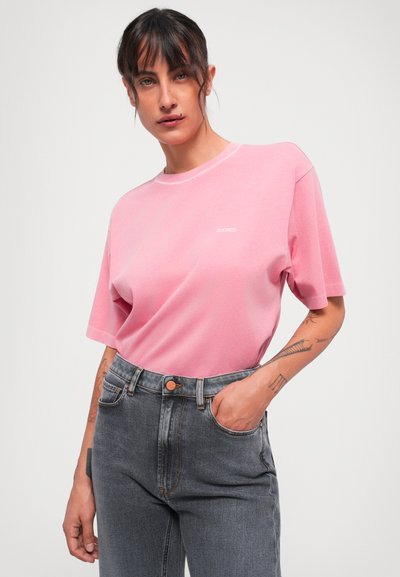 Woman with dark hair wearing a pink t-shirt with "CLOSED" text and high-waisted grey jeans, posing with one hand in pocket against white background.