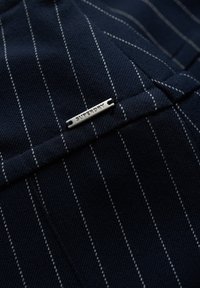 Tessuto blu navy con righe bianche, caratterizzato da un'etichetta in metallo argentato con la scritta "SUPERDRY" cucita nel design. Dettaglio della texture nitida visibile.