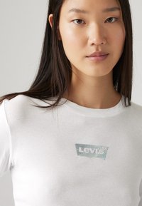 Valge puuvillane T-särk, millel on holograafiline Levi's logo rinnas, ümar kaelus ja lühikesed varrukad, sile tekstuur.