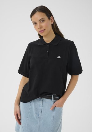 Polo noir à manches courtes, avec un col et un détail de logo blanc sur la côté gauche de la poitrine. Le modèle l'associe à une jupe en denim claire.