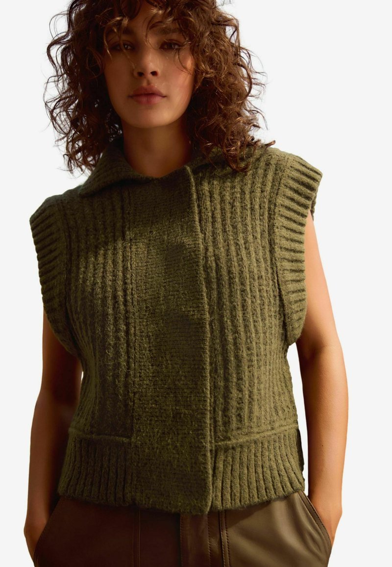 Gilet en tricot sans manches couleur kaki avec col montant, présentant une texture en côtes verticales et deux poches avant, réalisé en matériau doux et épais.