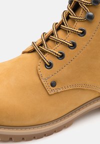 Bottine en suede beige avec œillets en métal noir, lacets bicolores et couture contrastante. Présente une semelle en caoutchouc avec un relief pour une meilleure adhérence.