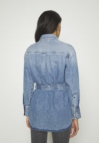 Denim srajca, svetlo modra, z pasom, z ovratnikom in z gumbi na manšetah. Mehka tekstura, vidne šivanke in subtilno zbledela.