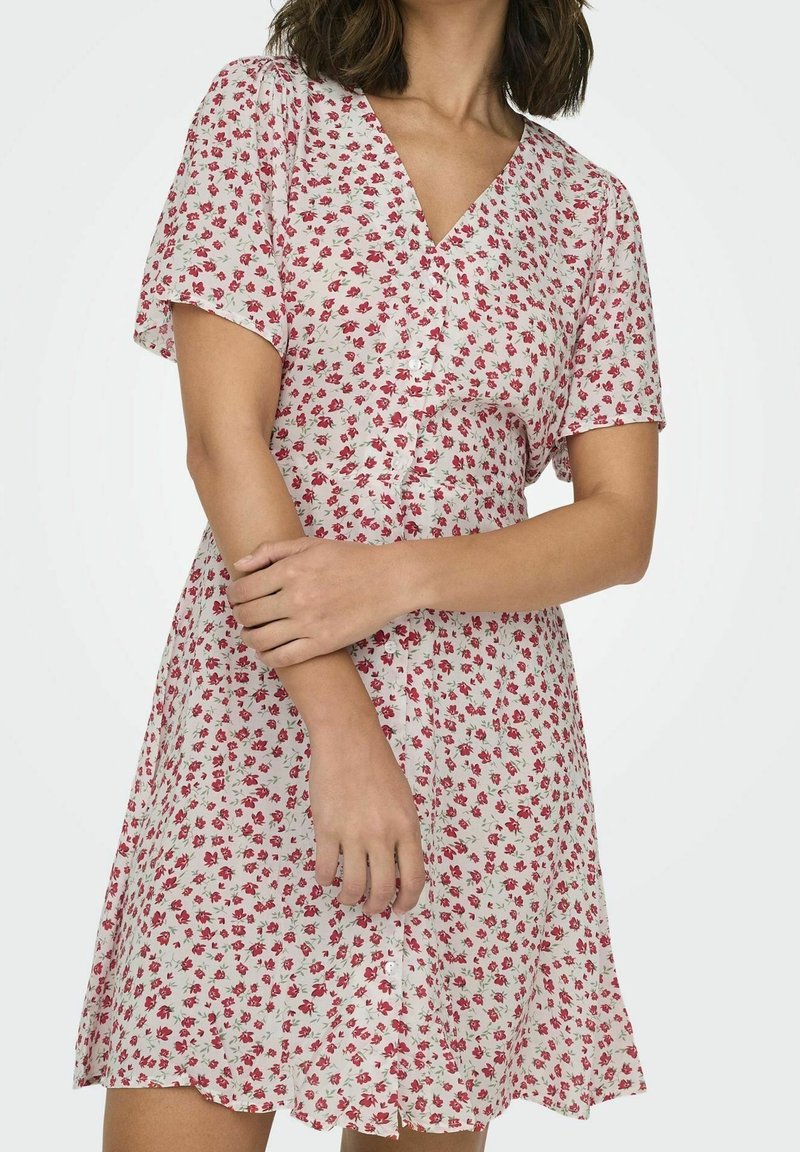 Femme portant une robe blanche à manches courtes avec un motif floral rouge, boutonnée à l'avant, avec un col en V, bras croisés à la taille.