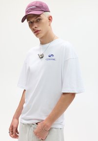 Hvid bomulds T-shirt med korte ærmer, der har blå grafisk tekst. Hudfarvet smykker og en burgunder kasket med teksten "EVER QUIET".