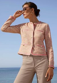 Rosa und cremefarbener Tweed-Blazer mit goldenen Knöpfen, gewellten Kanten und zwei Fronttaschen, kombiniert mit beigen Hosen vor einem blauen Ozeanhorizont.
