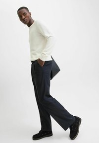 Chemise à rayures blanches à manches longues associée à un pantalon noir marine et des chaussures en suède noires. Le modèle se tient avec un sac noir sous le bras.
