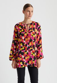 Langärmlige Bluse aus transparenter, floral gemusterter Stoff in den Farben Rosa, Gelb und Schwarz. Verfügt über einen Schlüssel-Ausschnitt mit einer Bindeschleife.