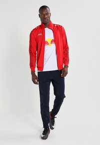 Rode sportieve jas met witte accenten en branding, gecombineerd met een witte t-shirt met een logo, marineblauwe broek en zwarte voetbalschoenen.