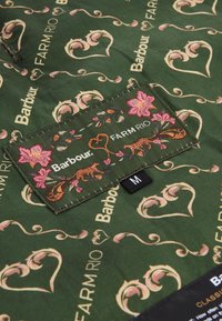 Πράσινο ύφασμα με σχέδιο που περιλαμβάνει διακοσμητικές καρδιές, ανθισμένα στοιχεία και κείμενο εμπορικής σήμανσης. Περιλαμβάνει ετικέτα με "Barbour," "FARM RIO," και μέγεθος "M."