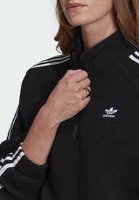 Černá fleece mikina s polovičním zipem a bílými třemi pruhy na rukávech, s vyšitým bílým logem. Měkký materiál, volný střih a stojáček.