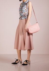 Blouse sans manches à imprimé floral avec un détail nœud, associée à une jupe midi plissée rose. Chaussures à talons carrés noires et argentées ; sac à main rose pâle.