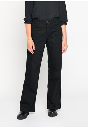 Person trägt schwarze, hoch taillierte Jeans mit geradem Bein, schwarzes langärmeliges Oberteil mit gerafften Manschetten und schwarze klobige Stiefel, steht vor weißem Hintergrund.