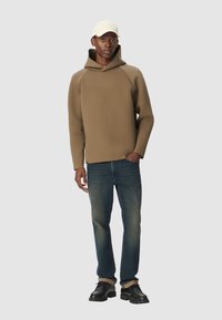 Mężczyzna ubrany w beżowy hoodie, niebieskie dżinsy, czarne buty i jasnobeżową czapkę, stojący z jedną ręką w kieszeni na jednolitym tle.