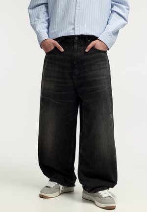 Personne portant un jean noir ample et délavé, des baskets grises avec des lacets blancs, et une chemise rayée bleu clair boutonnée, les mains dans les poches.