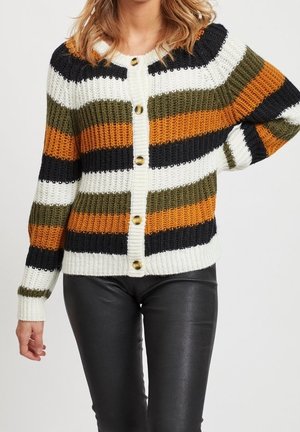 Donna che indossa un cardigan a coste abbottonato con strisce orizzontali in bianco, nero, verde oliva e arancione abbinato a pantaloni di pelle neri.