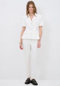 Witte op maat gemaakte blazer met korte mouwen, knopen en zakken, gecombineerd met bijpassende slanke broek en zwarte loafers met gouden accenten.