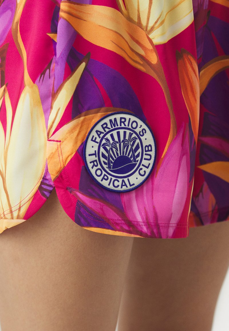 Jasno wzorzysty materiał zdobią tropikalne kwiaty w odcieniach różu, purpury i pomarańczu. Okrągła naszywka z napisem "FARMRIO'S TROPICAL CLUB" z grafiką palm.