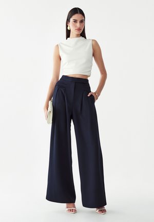 Femme portant un top court blanc sans manches, un pantalon large noir taille haute, des sandales blanches, tenant une pochette claire.