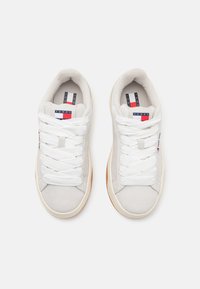 Baskets en suède gris clair avec des lacets blancs, semelle en gomme et un logo rouge et bleu bien visible sur la languette. Tige texturée au design minimaliste.