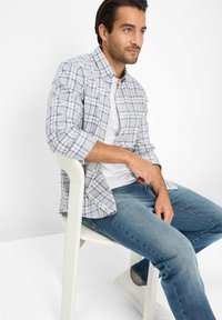 Lichtblauw overhemd met een ruitpatroon, met opgerolde mouwen en een borstzak, gedragen over een wit t-shirt en blauwe spijkerbroek.