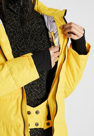 Doudoune jaune avec une fermeture éclair à deux voies, intérieur doublé et poche cachée. Haut noir en dessous avec un motif géométrique blanc.