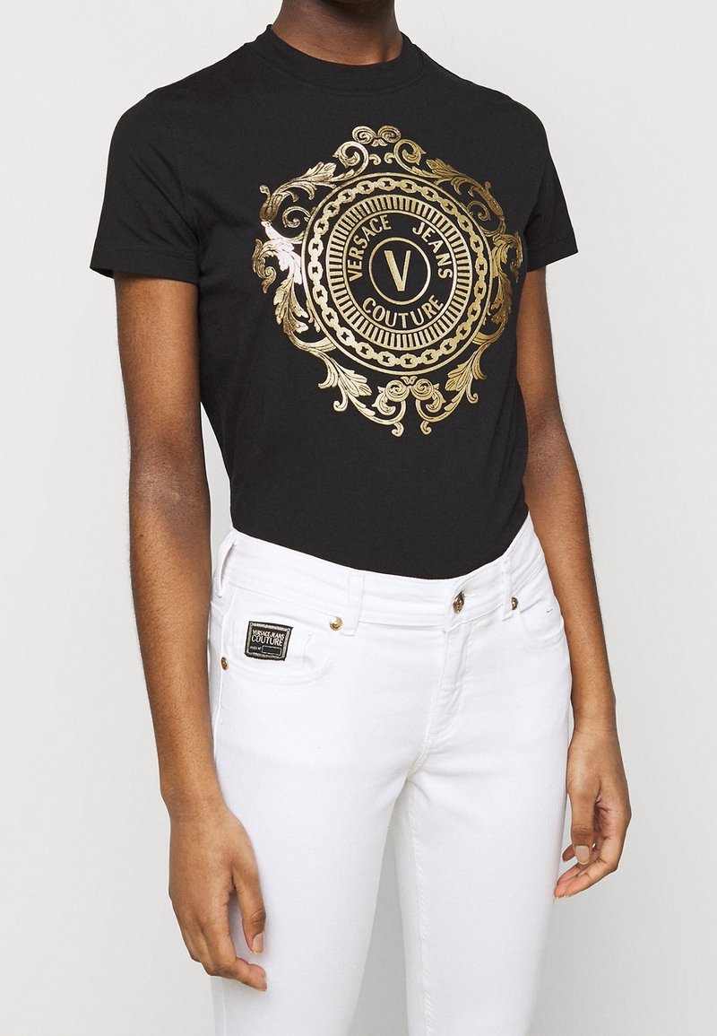 Musta t-paita, jossa on suuri kultainen barokki-tyylinen graafinen kuvio, jossa lukee "Versace Jeans Couture". Yhdistetty valkoisiin farkkuihin ja logo-tägeihin.
