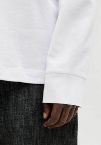 Hvid skjorte med lange ærmer og ribkant, vist sammen med mørkt denim, der fremhæver en glat tekstur og rene sømdetaljer.