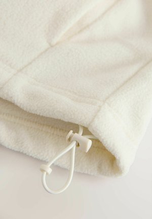 Vilnas apģērbs krēmkrāsā ar mīkstu tekstūru, tam ir aizdares poga un aukla apakšmalas regulēšanai.