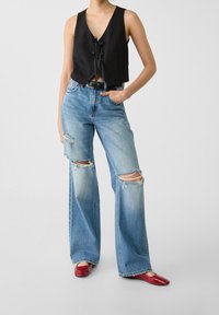 Black cropped vest with a tie front, high-waisted light blue distressed wide-leg jeans, and red flats with a glossy finish.