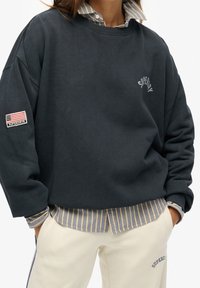 Osoba ubrana w ciemnogranatowy sweatshirt z logo Superdry, w paskowanym koszuli z guzikami pod spodem oraz w kremowe spodnie z napisem Superdry przy kieszeni.