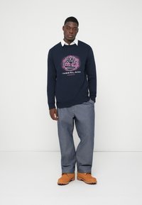 Bărbat îmbrăcat cu un Hanorac Timberland bleumarin, cămașă albă cu guler, pantaloni gri și cizme de lucru bej, stând în fața unui fundal alb simplu.