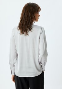 Lichtgrijze blouse met lange mouwen, gladde textuur, iets losse pasvorm, gegatherde manchetten en een gebogen zoom, gecombineerd met zwarte broek.