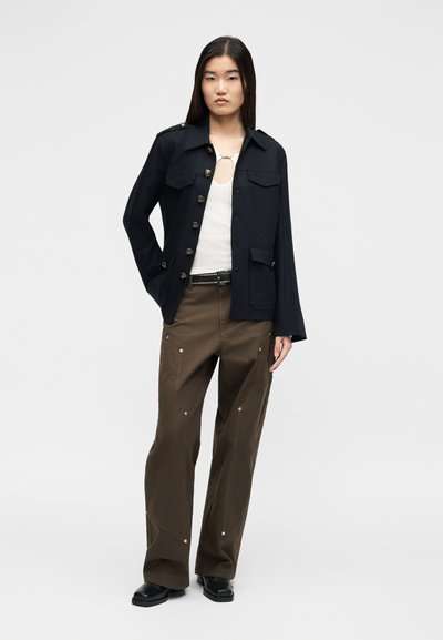 Γυναίκα στέκεται φορώντας μαύρο σακάκι με κουμπιά, λευκό τοπ με άνοιγμα, καφέ φαρδιά παντελόνια, μαύρα παπούτσια και μαύρη ζώνη με τρουκς σε απλό φόντο.