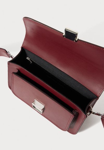 Τσάντα χειρός από δέρμα μπορντό με ασημένιο κούμπωμα και εσωτερική τσέπη με φερμουάρ. Διαθέτει υφή επένδυσης και ρυθμιζόμενο λουρί.