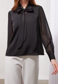 Kvinna som bär en svart satinblus med genomskinliga långärmade prickar och en knuten slipskrage, tillsammans med vita byxor.