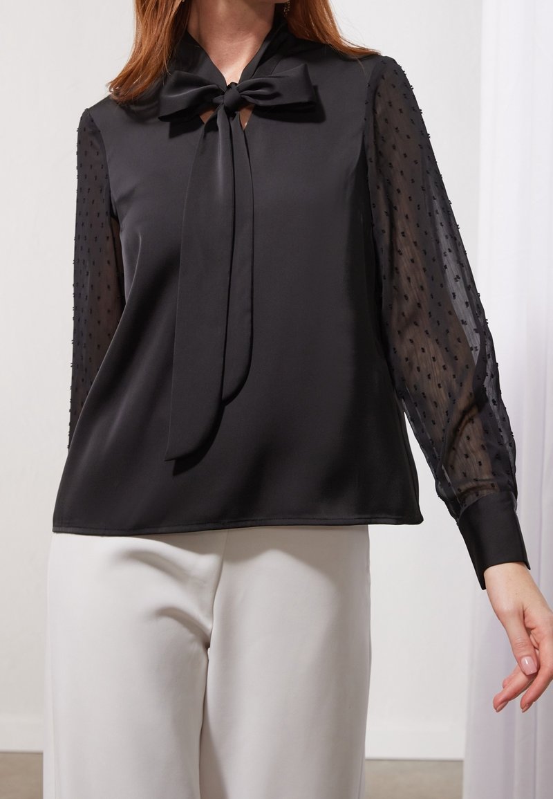 Kvinna som bär en svart satinblus med genomskinliga långärmade prickar och en knuten slipskrage, tillsammans med vita byxor.