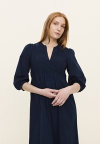 Navyblaues Kleid aus einem Stoff mit strukturiertem Smock-Oberteil, einem V-Ausschnitt und dreiviertellangen Puffärmeln.