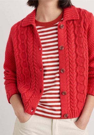 Person trägt eine rote, kabelgestrickte, geknöpfte Strickjacke über einem rot-weiß gestreiften Hemd, die Hände in den hellfarbenen Hosentaschen.