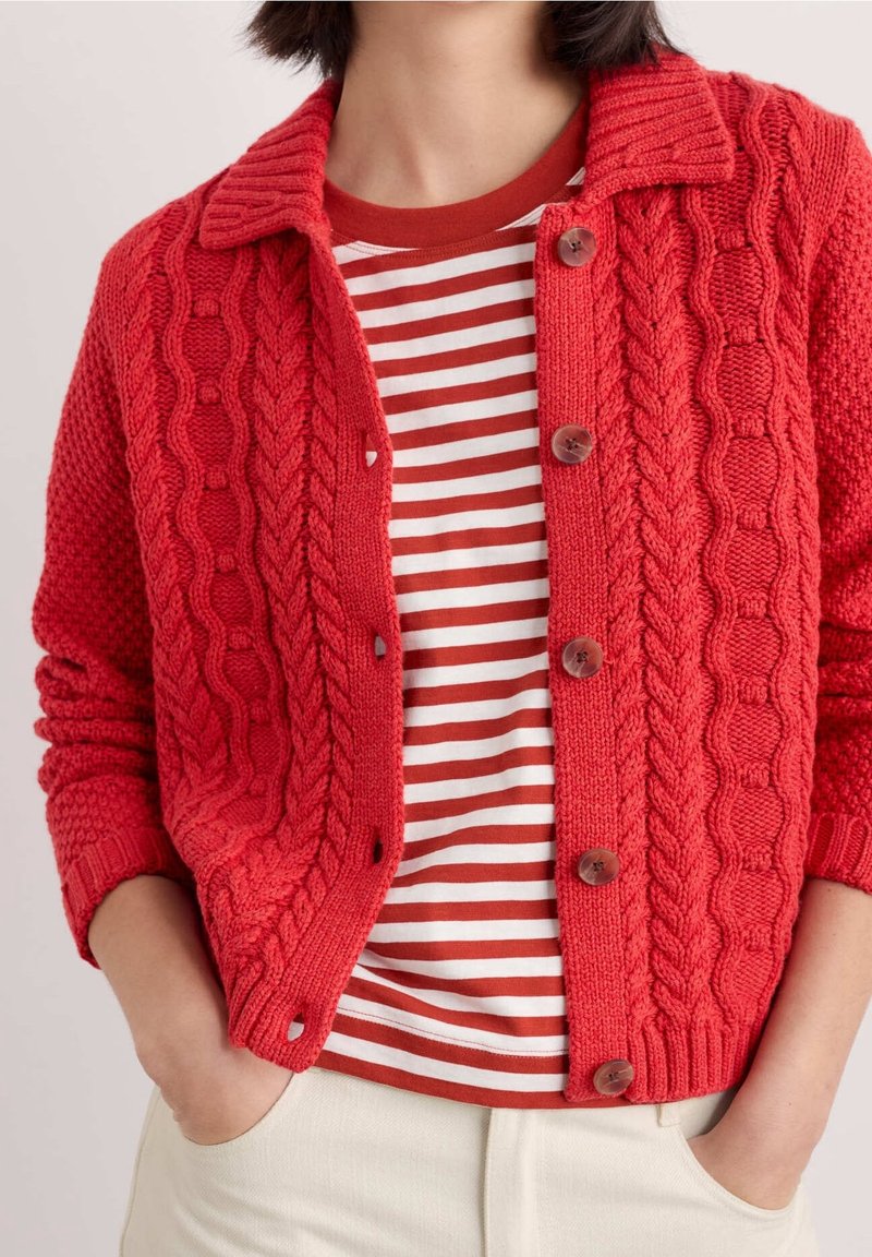 Personne portant un cardigan rouge en maille torsadée boutonné sur une chemise rayée rouge et blanche, les mains dans les poches d'un pantalon clair.
