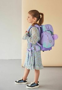 Lila und türkisfarbener Rucksack mit einem flauschigen Pompon, gepolsterten Trägern und mehreren Fächern. Blümchenmusterkleid und schwarze Sneaker getragen.