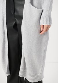 Hellgrauer Cardigan aus weichem Strickstoff, mit gerippten Bündchen und seitlichen Taschen, getragen über schwarzen Lederhosen.