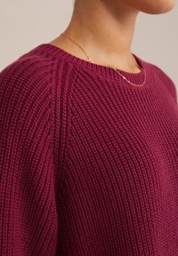 Stickad tröja i djup burgunder, med rund hals och texturerat ribbmönster. Tyget ser mjukt ut med synliga sömnadsdetaljer.